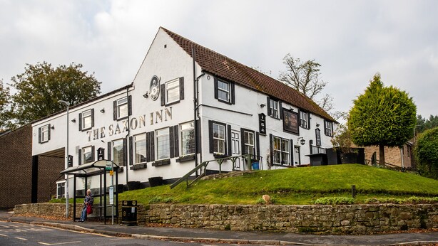 A traditional building with a sign reading "The Saxon Inn." It features a stone wall, a grassy hill, and a paved road in front. - The Saxon Inn (Bishop Auckland)