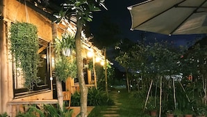 A well-lit outdoor area with a pathway leading to a building, surrounded by greenery and potted plants.