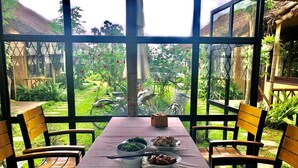 A dining area with a table set for two, surrounded by chairs, overlooking a garden with a bicycle and a thatched roof structure.