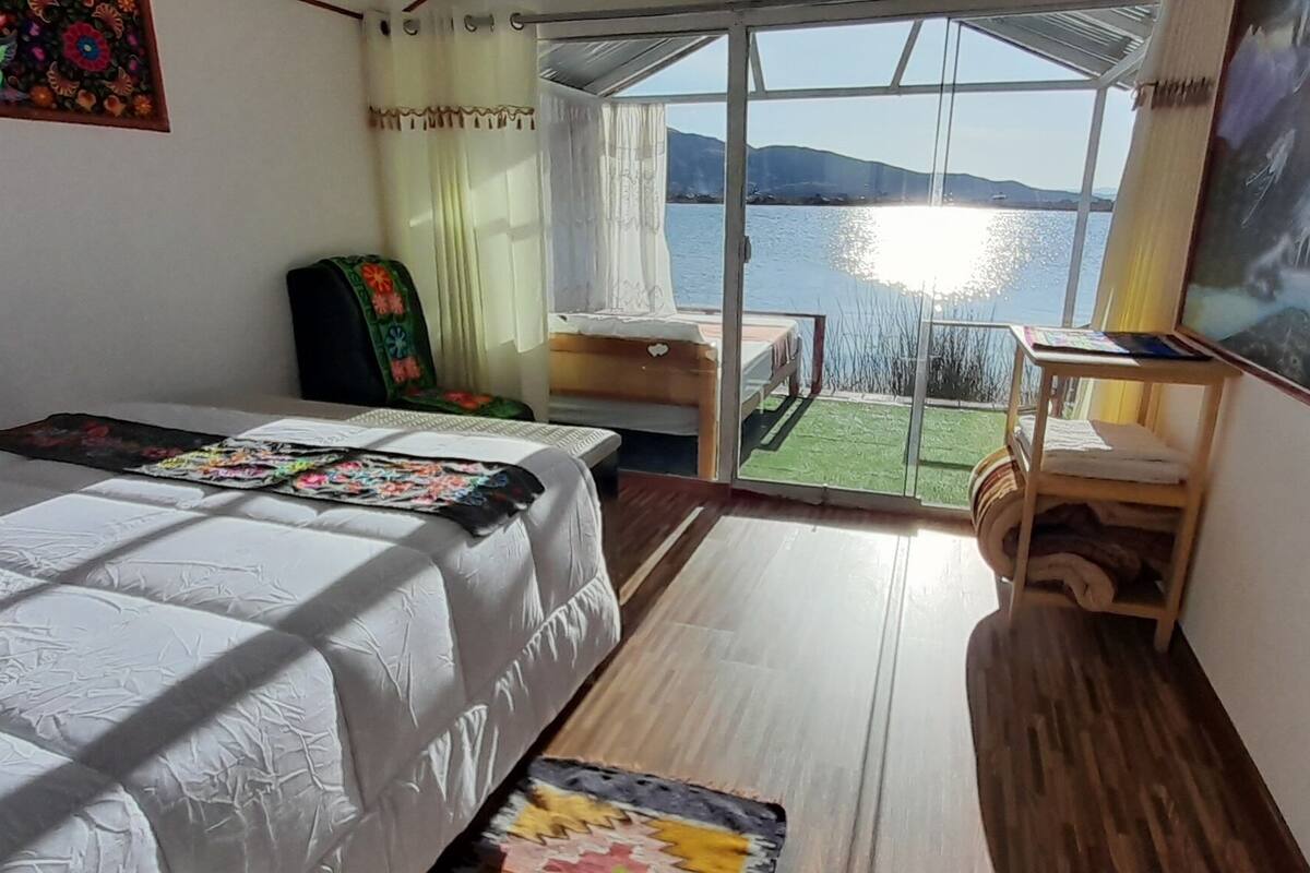 A bedroom with a bed, a small table, a chair, a window with a view of the sea, and a painting on the wall.