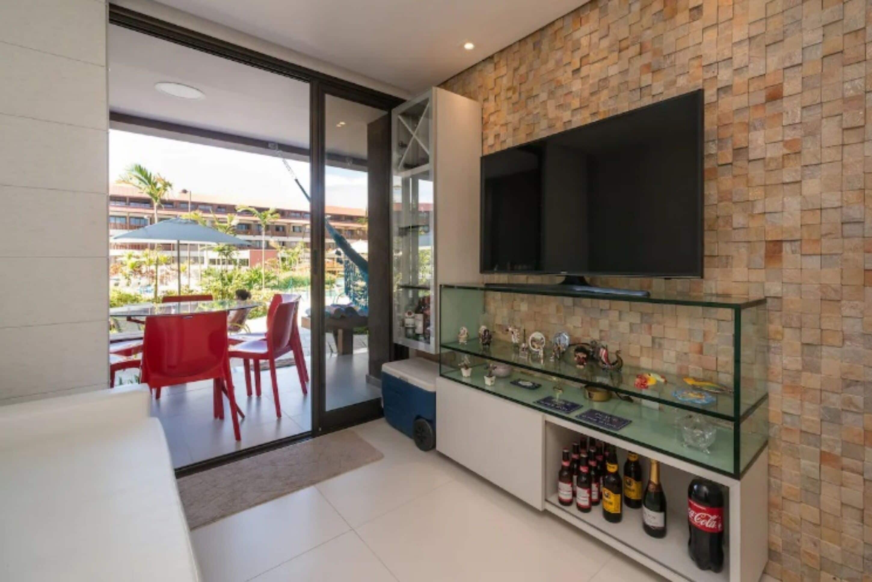 A modern hotel room with a large TV mounted on a brick wall, a glass display case with bottles, and a view of the outdoor pool area.