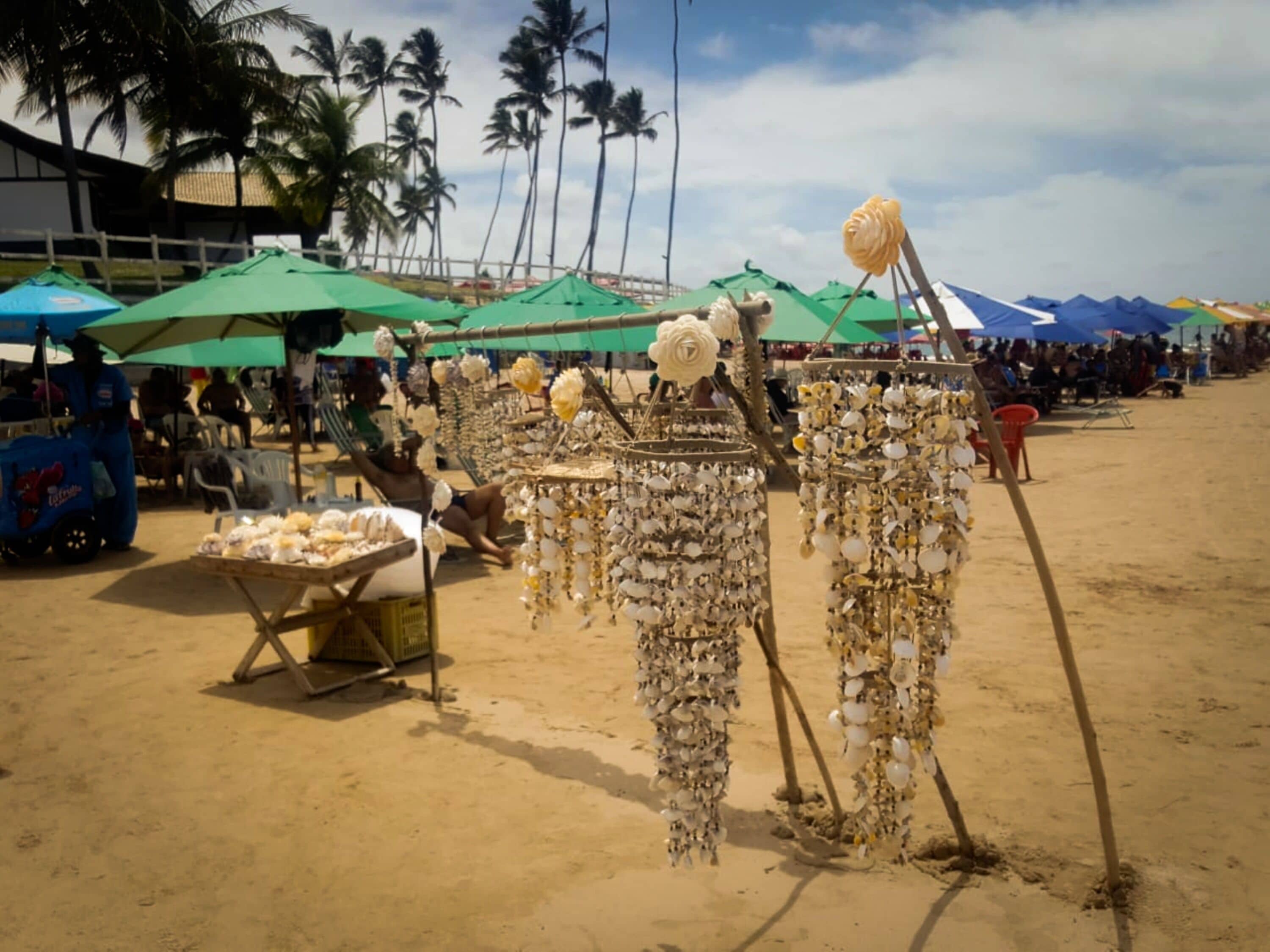 A beach market with stalls selling jewelry and souvenirs, surrounded by palm trees and beach umbrellas.