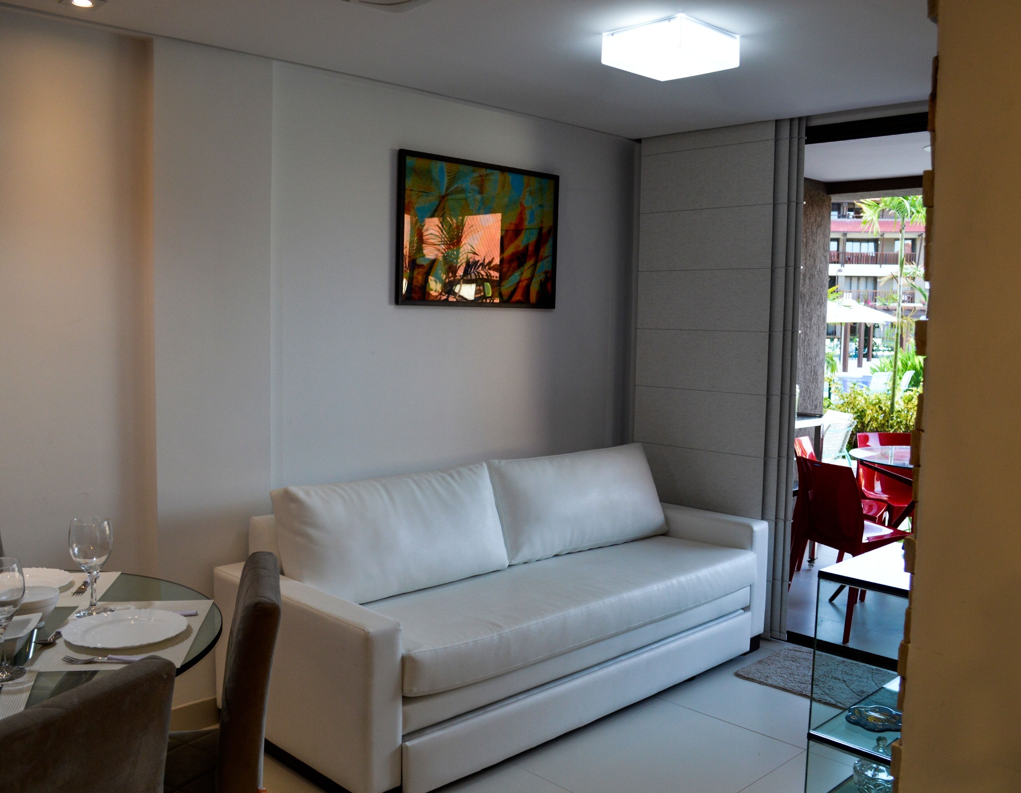 Uma sala de estar moderna com um sofá branco, uma mesa de jantar com cadeiras e uma mesa de centro de vidro. Há um quadro na parede e vista para a área do balcão.