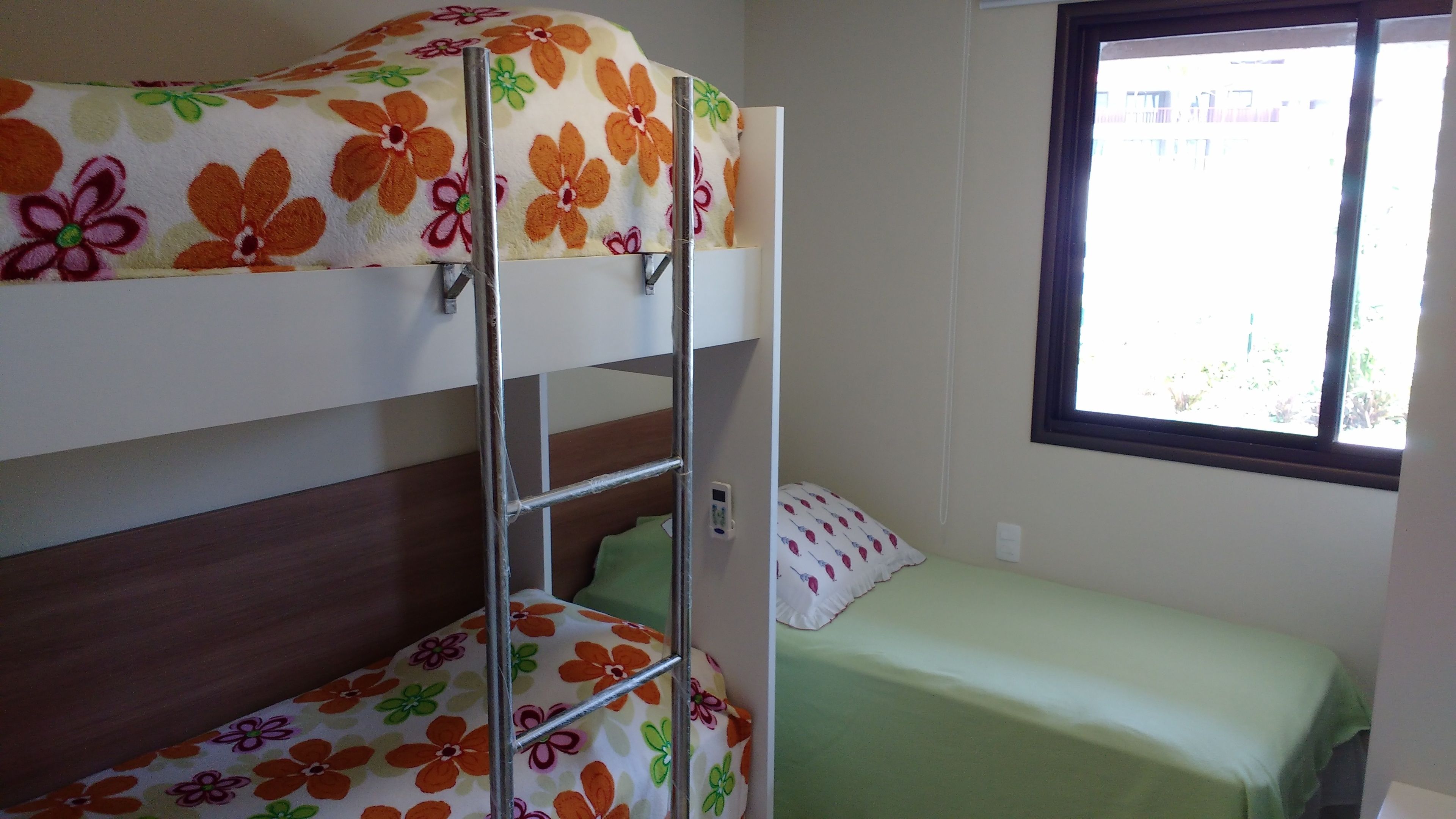 A bunk bed with floral bedding, a single bed with a green bedspread, and a window with a view of the outside.