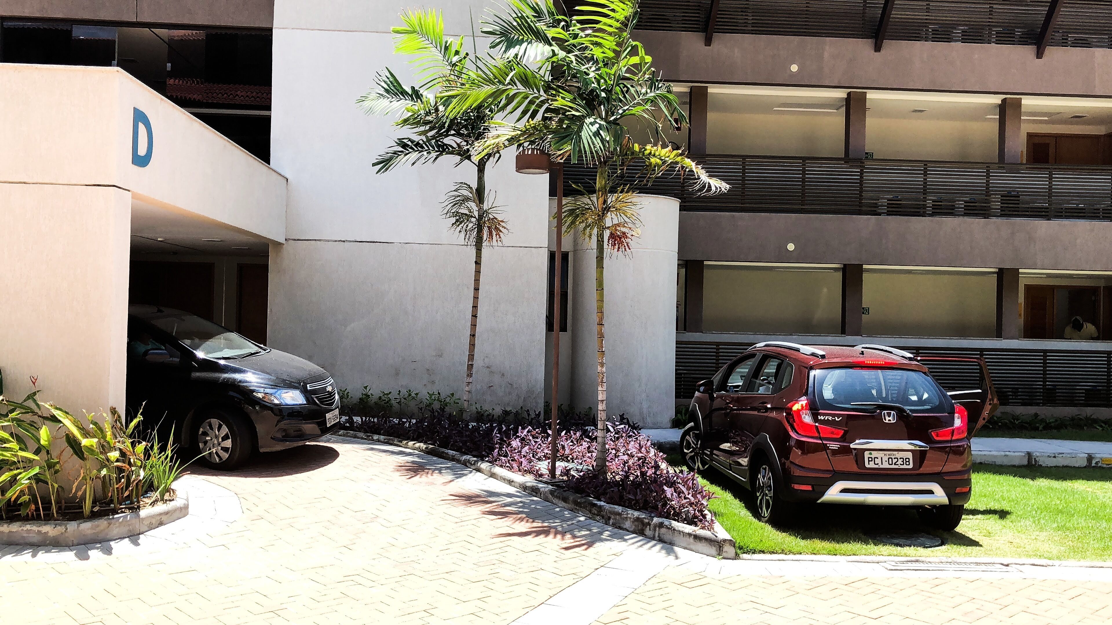 Ein Parkplatz mit zwei Autos, ein Gebäude mit einem ‚D‘-Schild und ein kleiner Garten mit Pflanzen.