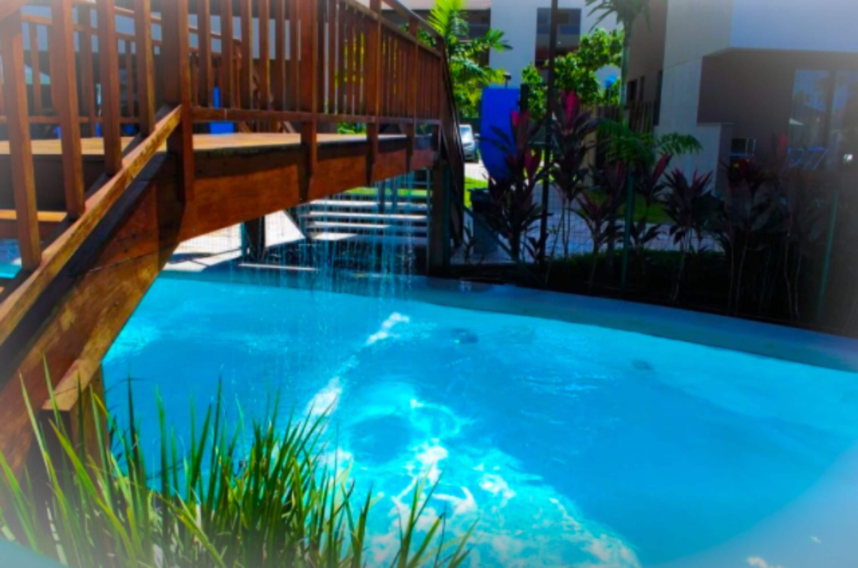 Piscina com uma ponte de madeira e escadas que a levam, rodeada por vegetação.