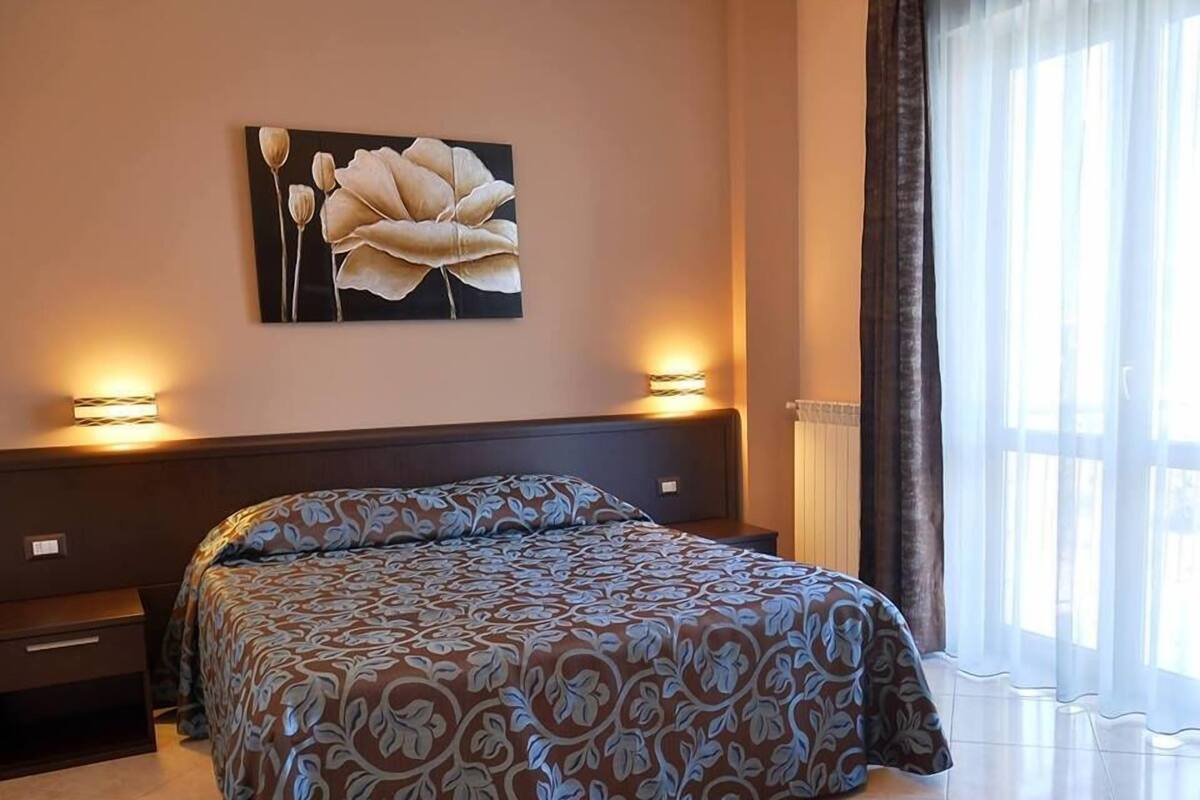 Ein Schlafzimmer mit einem großen Bett, zwei Nachttischen, einem Wandbild und einem Fenster mit Vorhängen.