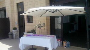 Un patio al aire libre con una mesa cubierta por un mantel blanco y un paraguas grande. Hay una planta en maceta sobre la mesa, y un bote de basura es visible a la izquierda.
