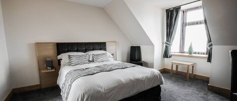 A bedroom with a large bed, a bedside table, a lamp, a window with curtains, and a small bench.