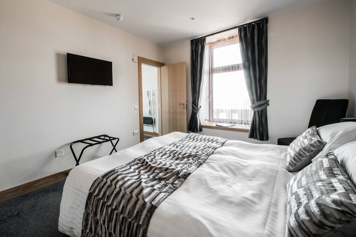 A hotel room with a bed, a TV mounted on the wall, a window with curtains, and a door.