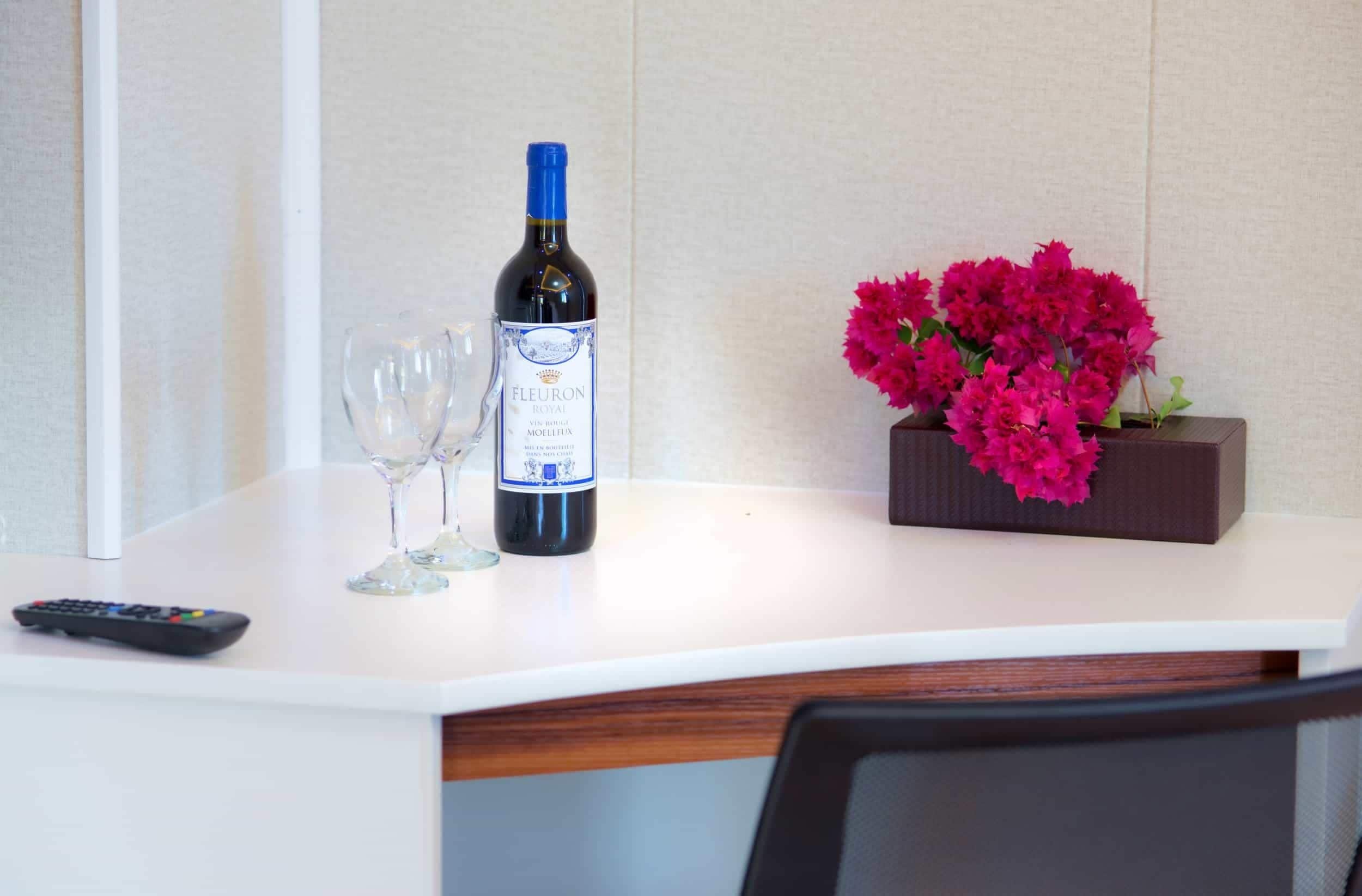 A hotel room with a white table, a bottle of wine, two wine glasses, and a vase of pink flowers.
