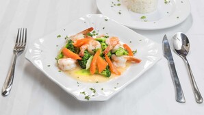 A square white plate with a seafood dish, a fork, a knife, and a spoon on a white tablecloth.