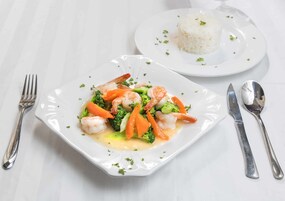 Un plato cuadrado blanco con un plato de mariscos, un tenedor, un cuchillo y una cuchara sobre un mantel blanco.