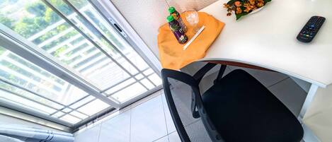 A room with a large window, a desk with a remote control, a bottle, a glass, and a flower arrangement.