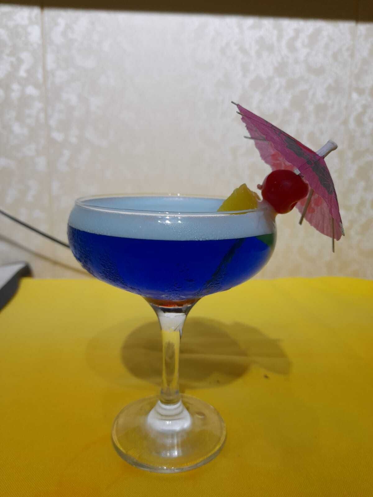 Un cóctel en vaso azul, con cereza y un paraguas decorativo sobre una mesa amarilla.
