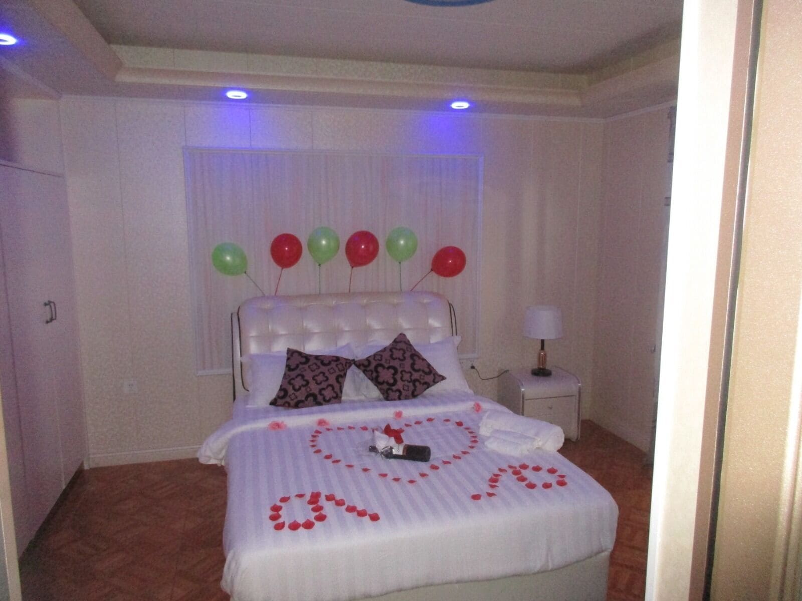A neatly made bed with a heart-shaped arrangement of rose petals and a small gift box, a white headboard with balloons, and a bedside table with a lamp.