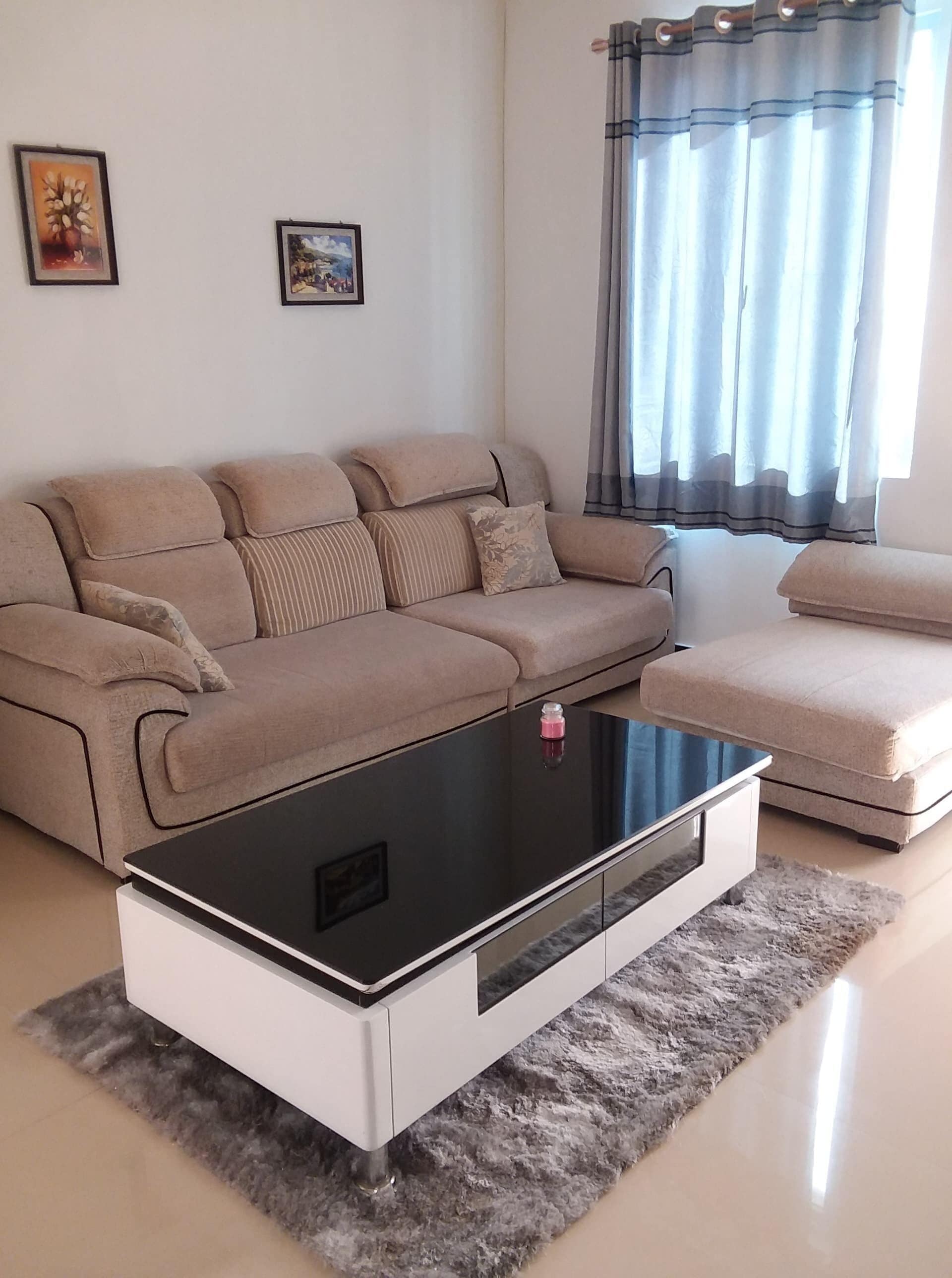 A living room with a beige sofa, a white armchair, and a black coffee table. There are two framed pictures on the wall.
