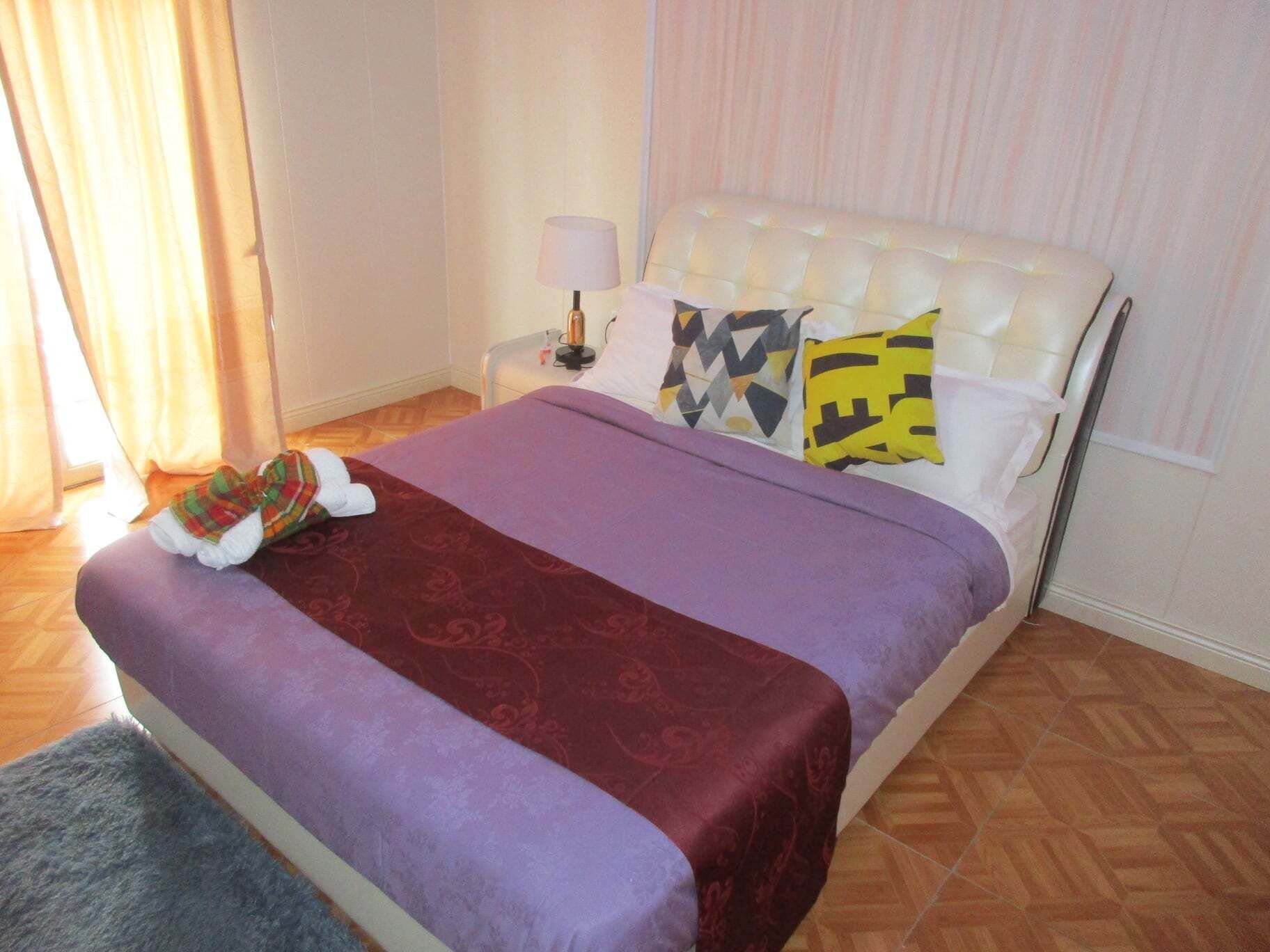 Un lit bien fait, avec une couvre-lit violette et une couverture rouge, une tête de lit blanche et des coussins à motifs.