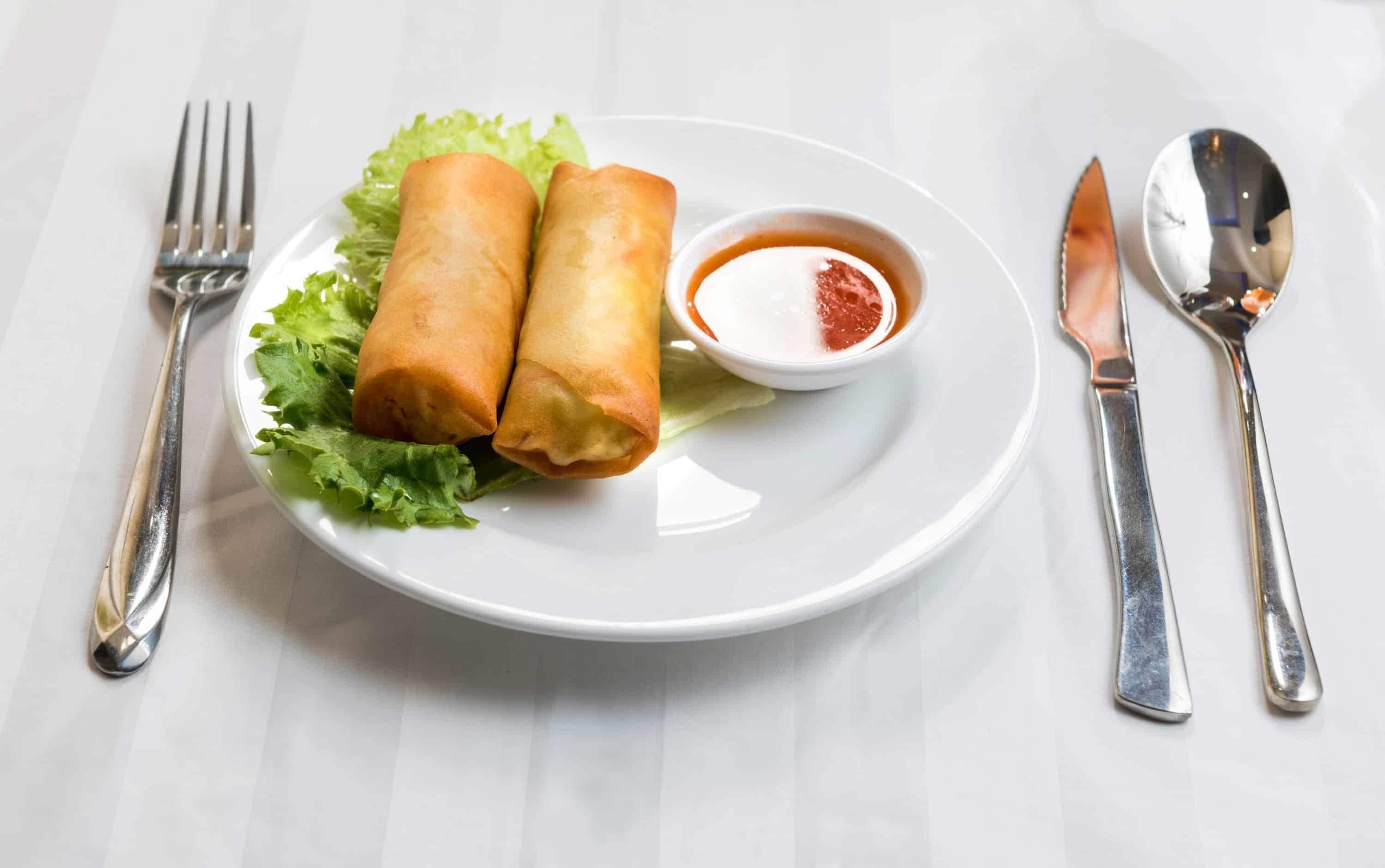 Un plato con dos rollitos primavera, un recipiente pequeño de salsa para mojar y cuchara y tenedor a un lado.