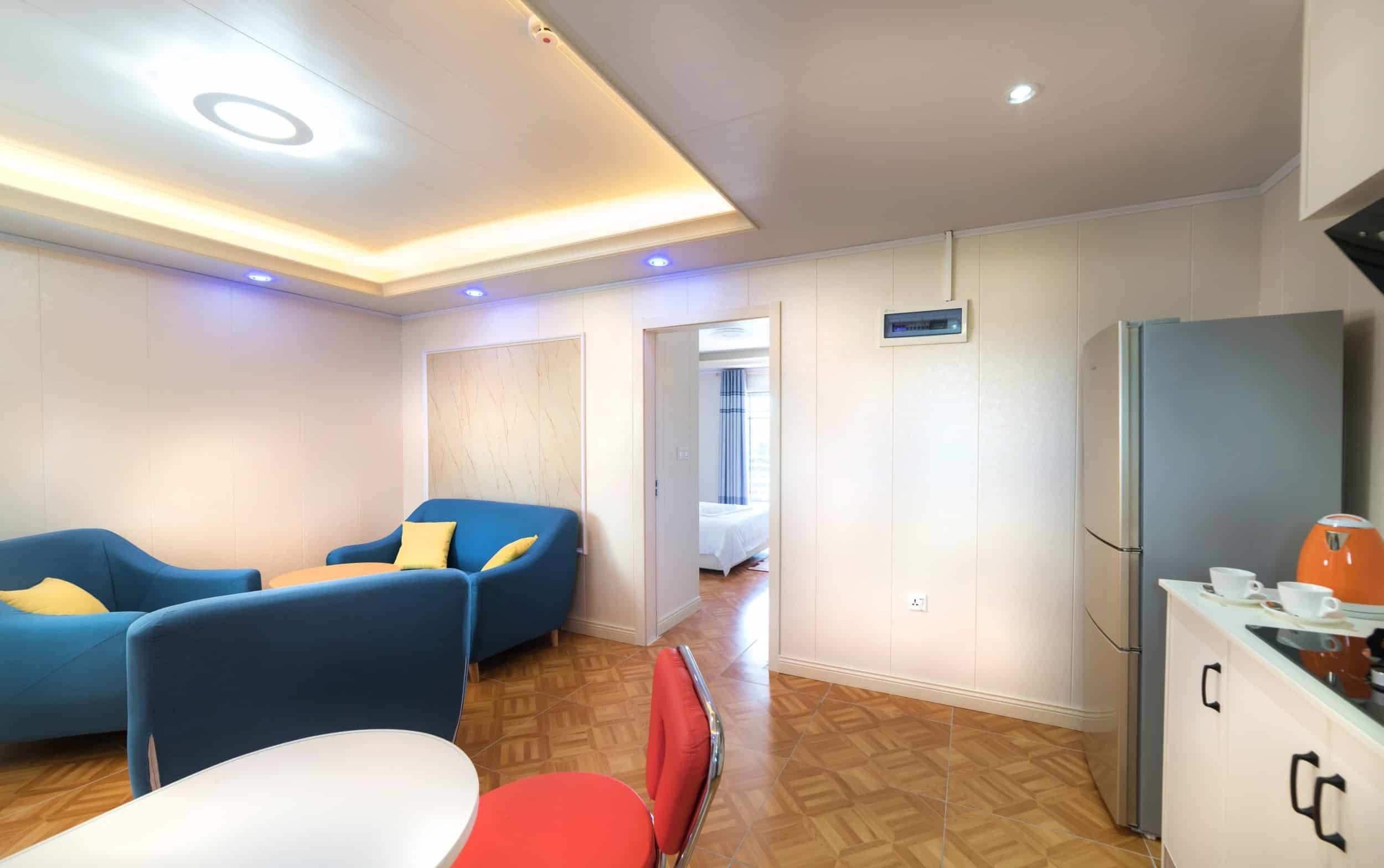 A modern hotel room with a wooden floor, a bed with a blue and white striped curtain, a blue sofa with yellow cushions, a red chair, a white table, and a kitchen area with a refrigerator and a kettle.