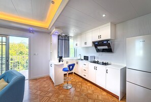 Una cocina moderna con armarios blancos, microondas integrado y refrigerador. Hay un taburete azul y una ventana con vista a la vegetación.