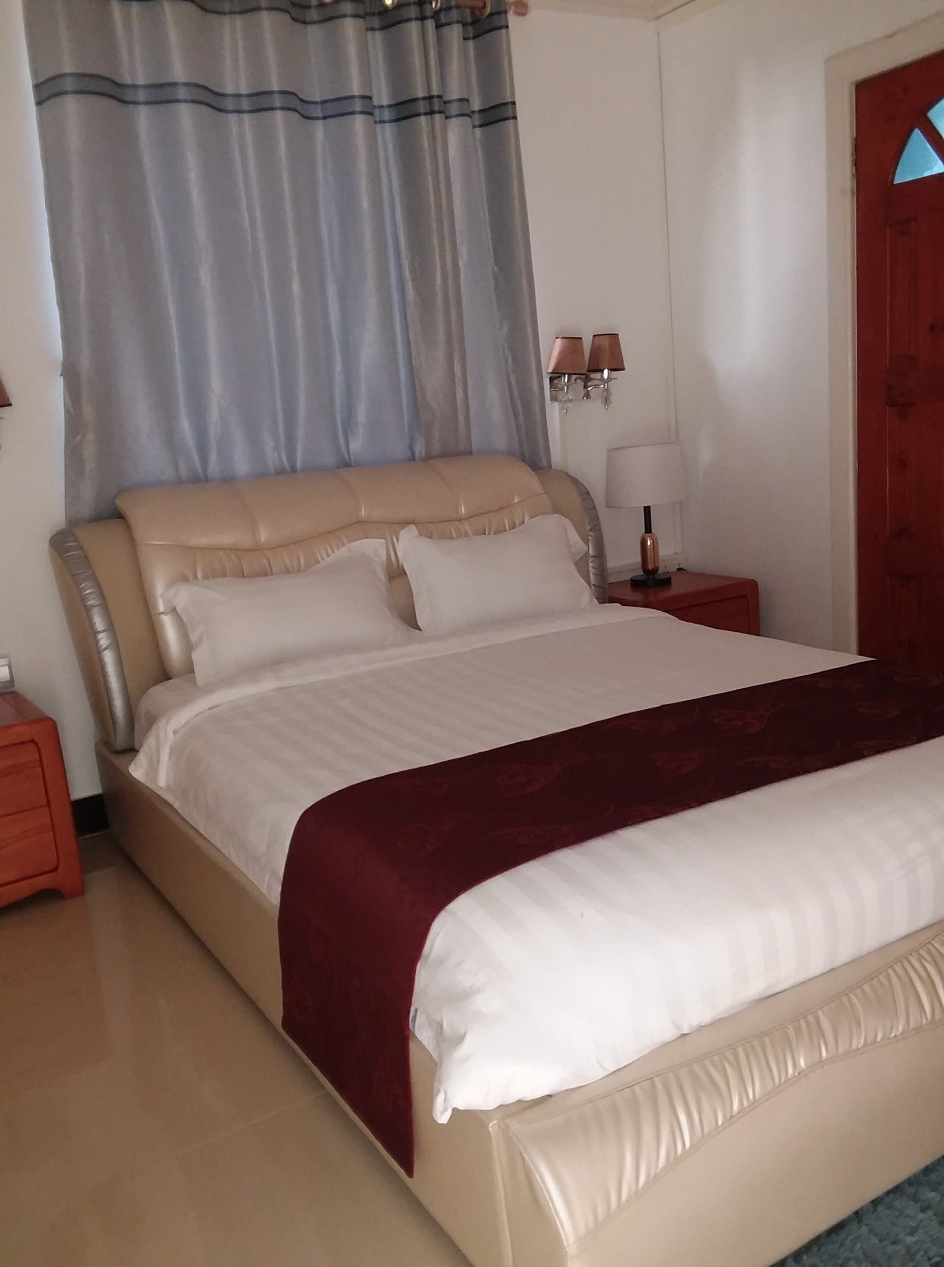 A neatly made bed with a beige headboard and white bedding, a red accent pillow, and a red throw blanket. There is a nightstand with a lamp and a wall-mounted light fixture.
