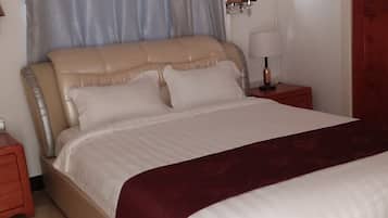 Een net opgemaakt bed met een beige hoofdbord en witte beddengoed, een rode kussen en een rode plaid. Er staat een nachtkastje met een lamp en een wandlamp.