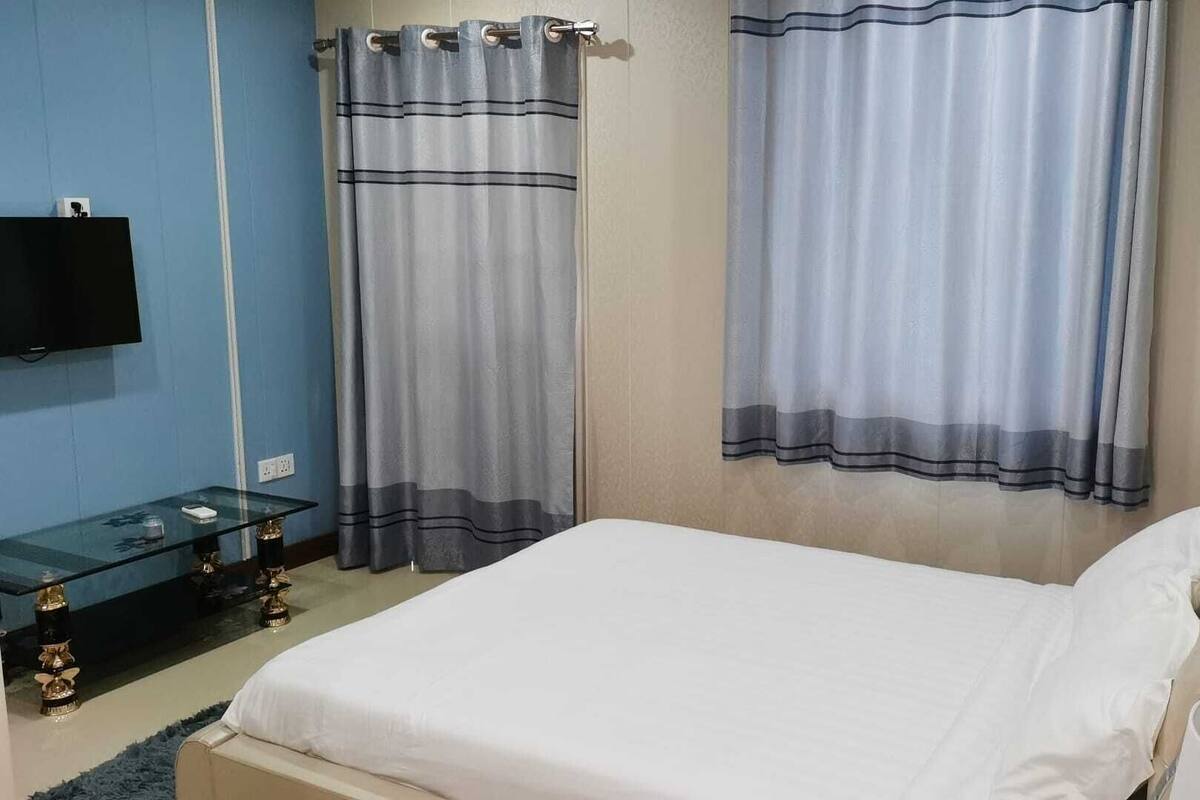 A bedroom with a bed, a glass side table, a TV mounted on the wall, and two curtains.