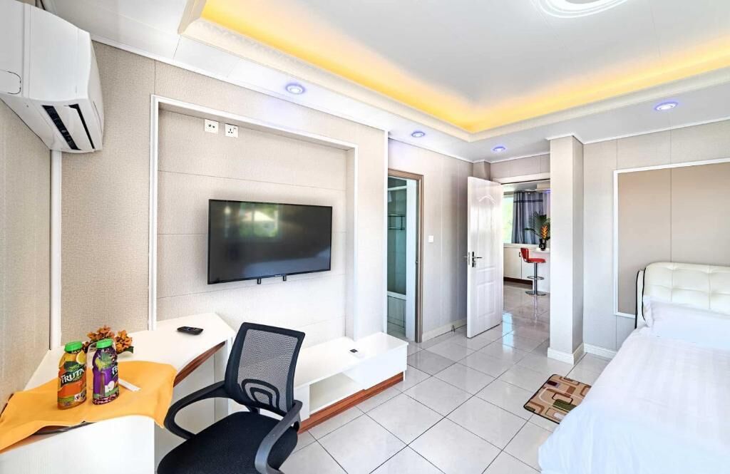 A hotel room with a bed, a desk with a chair, a TV mounted on the wall, and a door leading to another room.