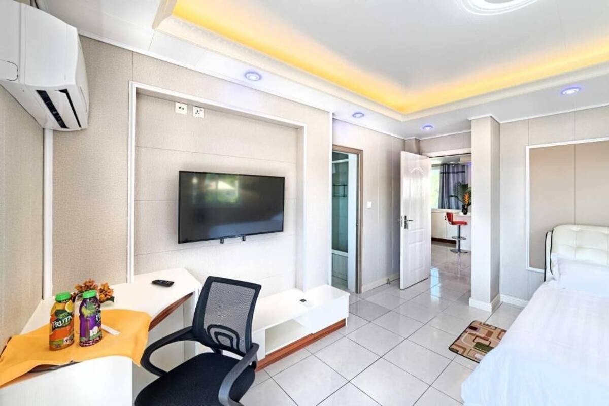 A hotel room with a bed, a desk with a chair, a TV mounted on the wall, and a door leading to another room.