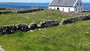 Una casa blanca con techo inclinado y tragaluces, rodeada por un muro de piedra y un campo cubierto de hierba, con vista al mar en el fondo.