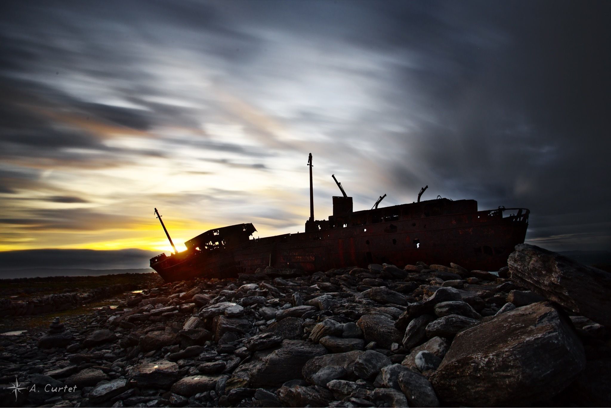 A shipwreck on a rocky shore with a sunset in the background.