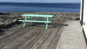 Una terraza de madera con una mesa de picnic, un camino de hormigón y una orilla rocosa con vista al océano.