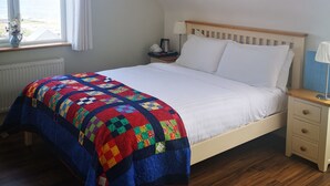 Un dormitorio con cama de madera, ropa de cama blanca, colcha colorida, mesita de noche con lámpara, ventana con cortinas y radiador.