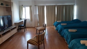 Ett hotellrum med trägolv, ett skrivbord med en stol, en tv och tre sängar med blått sänglinne.
