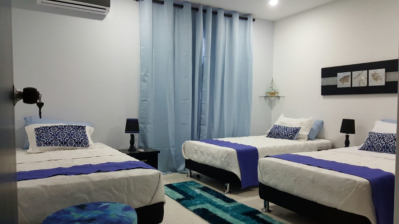 A hotel room with two beds, a nightstand, a lamp, a window with curtains, and a wall-mounted shelf with decorative items.