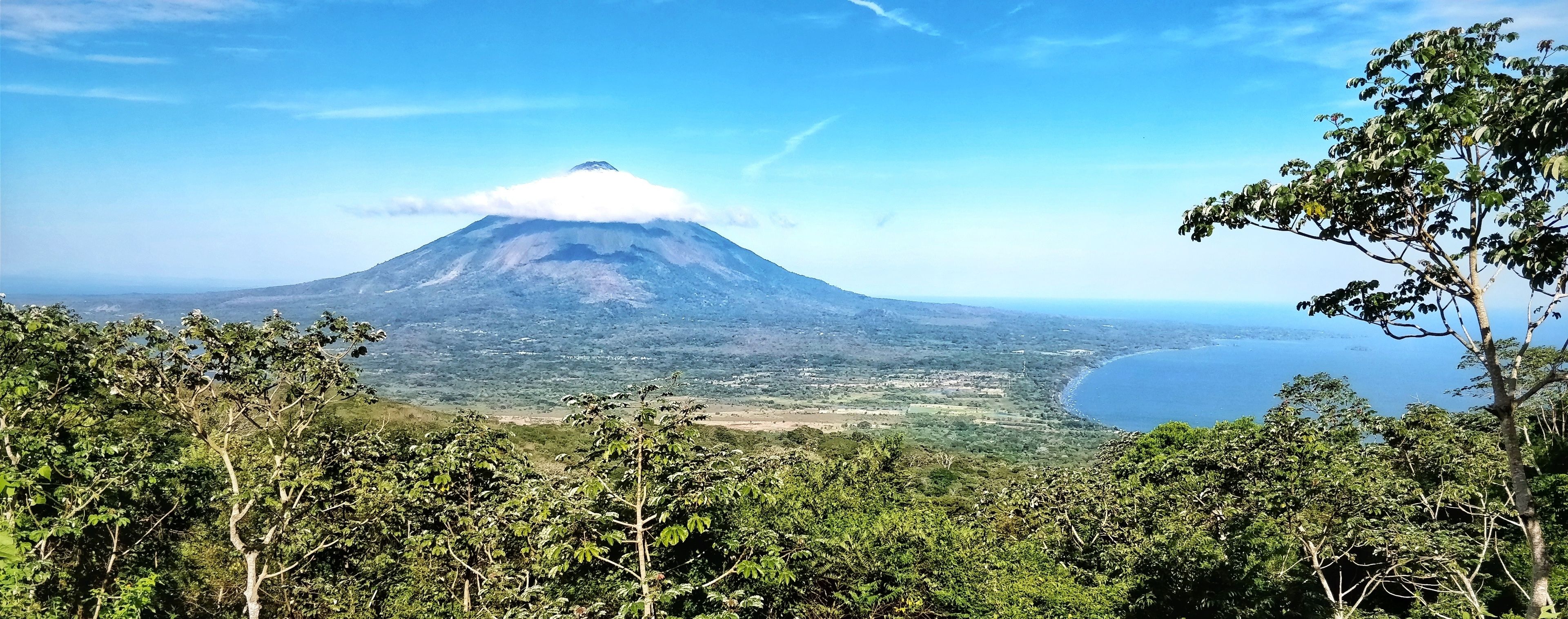 A mountain with a cloud base, surrounded by dense greenery and a coastal view.