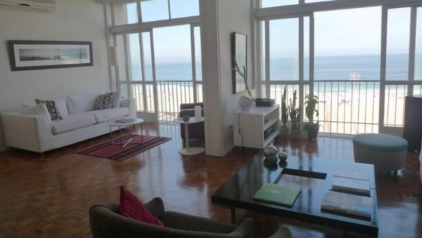 A spacious living room with a white sofa, a coffee table, and a view of the beach through large windows. - Vista Espetacular Copacabana (Rio de Janeiro)
