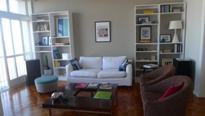 A living room with a white sofa, a brown armchair, a coffee table, and two bookshelves filled with books and decorative items. - Vista Espetacular Copacabana (Rio de Janeiro)