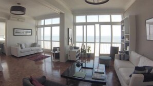 A spacious living room with a large window overlooking the sea, a white sofa, a coffee table, and a bookshelf. - Vista Espetacular Copacabana (Rio de Janeiro)