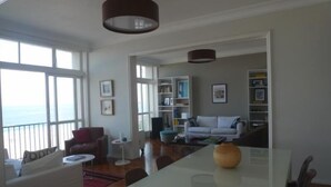 A spacious living area with a large window overlooking the sea, a white sofa, a red armchair, a white coffee table, and a dining table with a vase. - Vista Espetacular Copacabana (Rio de Janeiro)