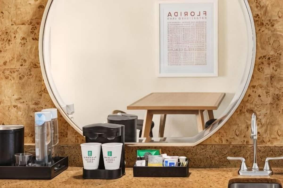 A hotel room with a round mirror, a wooden table, a sink, and various toiletries on the counter.