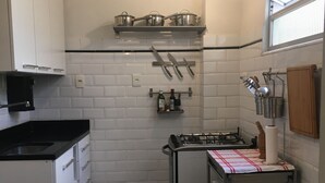 A compact kitchen with white cabinets, a gas stove, a small refrigerator, and a hanging pot rack. - Leblon 2 Quartos 500 Metros da Praia (Rio de Janeiro)