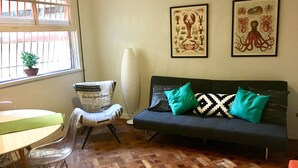 A living room with a wooden parquet floor, a black sofa with patterned cushions, a clear plastic chair, a table with a green tablecloth, a potted plant, and two framed wall art pieces. - Leblon 2 Quartos 500 Metros da Praia (Rio de Janeiro)