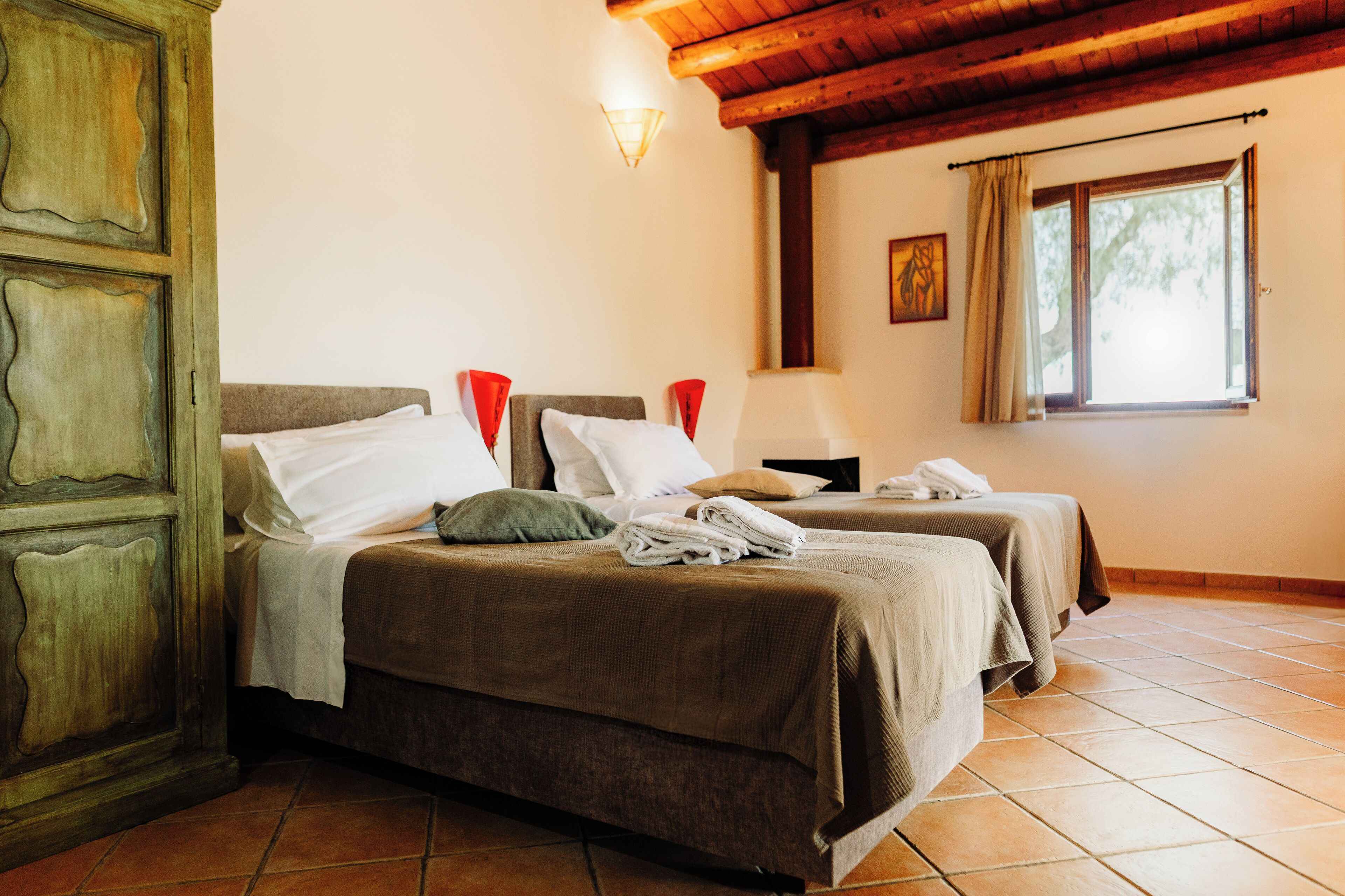 A room with two beds, a wooden wardrobe, a window with curtains, and a wall-mounted light fixture.
