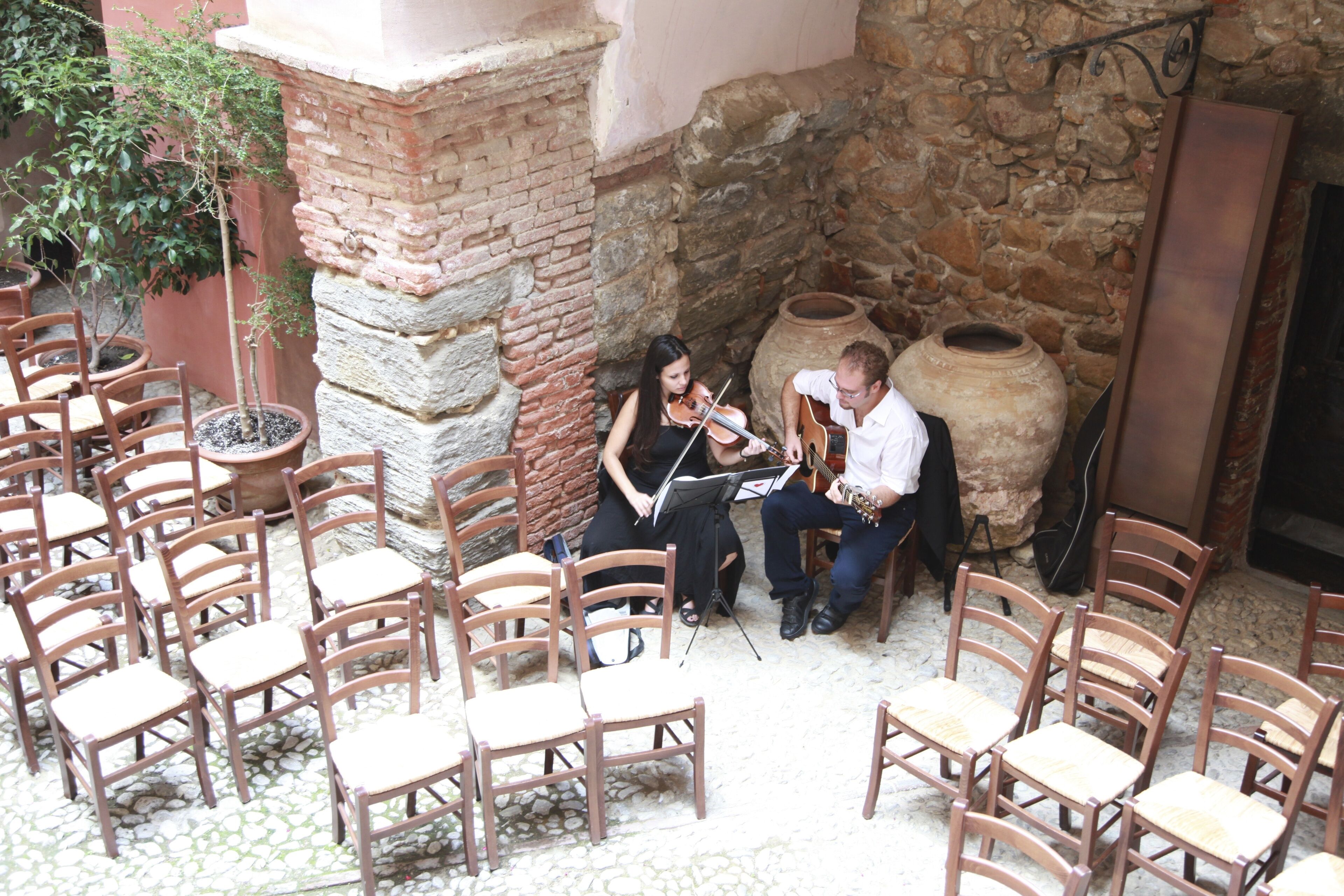 Une cour intérieure avec des chaises en bois disposées pour que les gens puissent s’asseoir, un violoniste et un guitariste qui jouent, et un mur de pierre en arrière-plan.