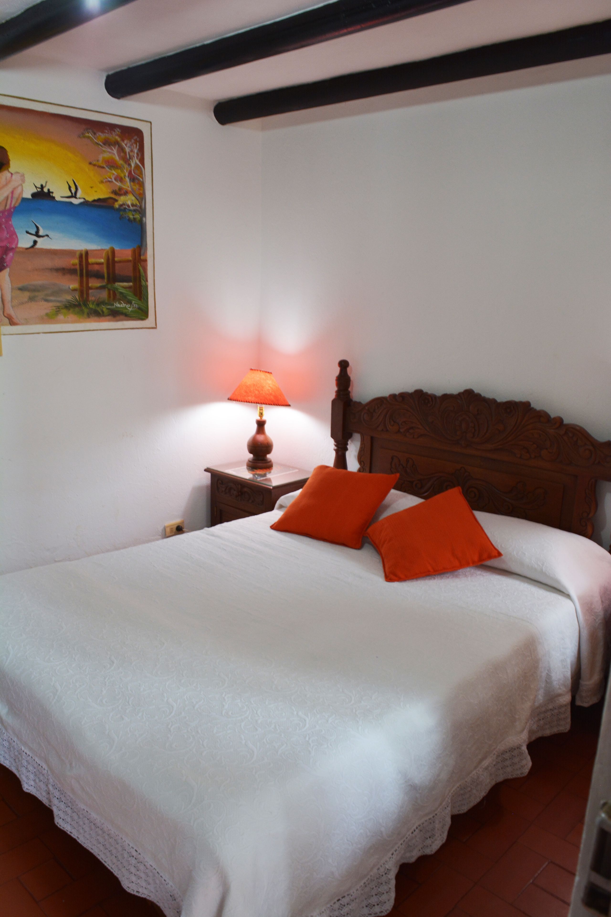 A bedroom with a wooden headboard, a white bedspread, two orange pillows, a nightstand with a lamp, and a painting on the wall.