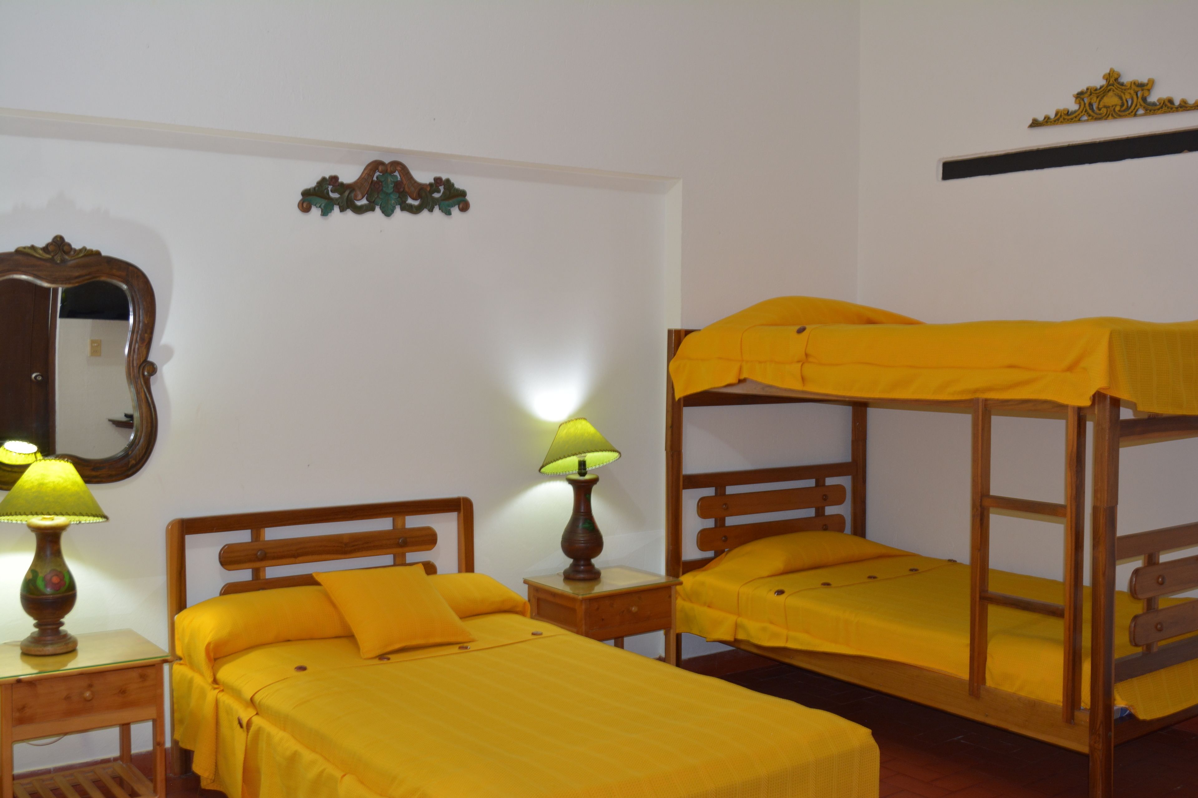 A room with two wooden bunk beds, a single bed, a nightstand with a lamp, a mirror, and a decorative wall piece.