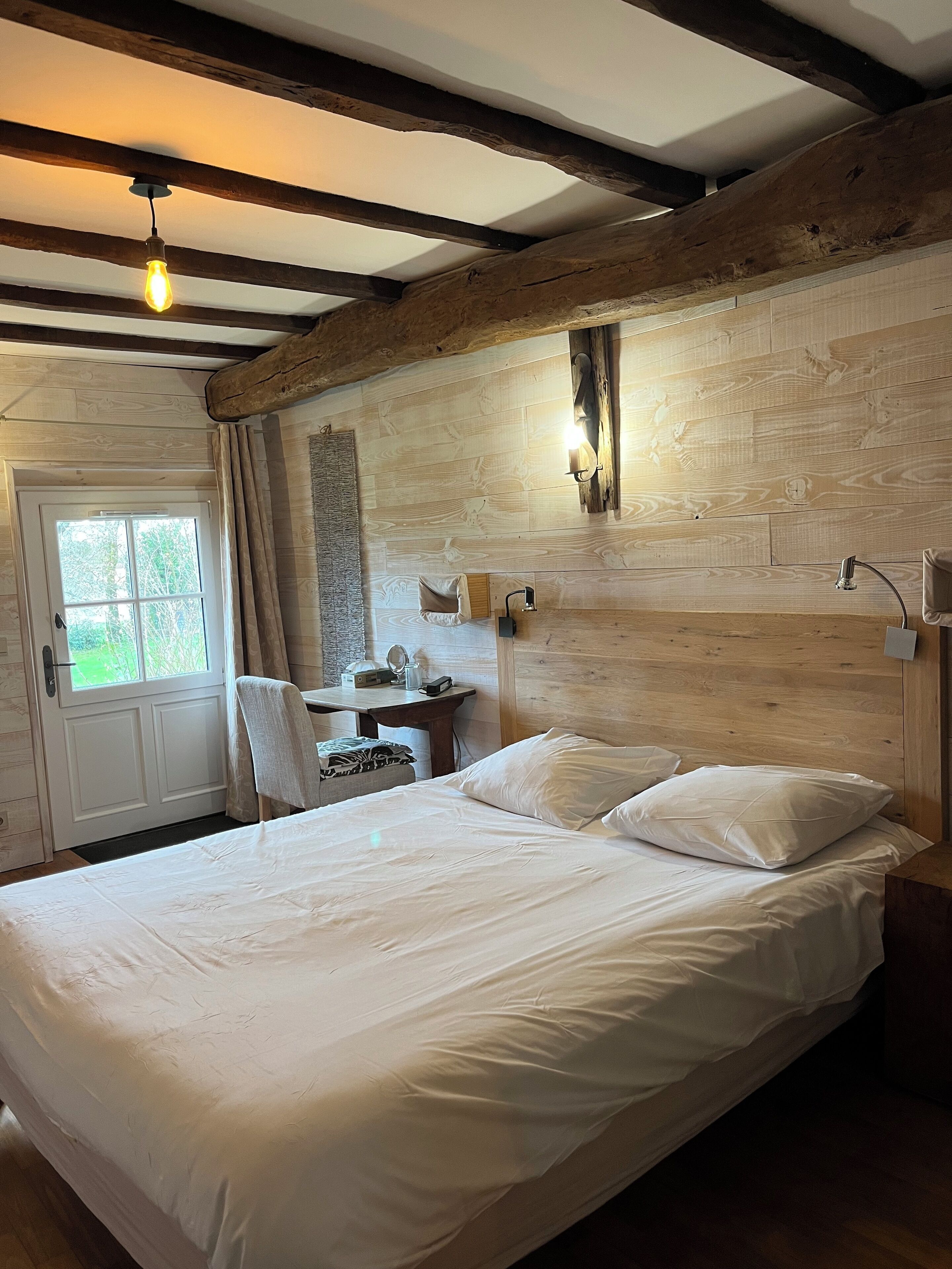Une chambre avec un grand lit, des poutres en bois, un bureau avec une chaise et une fenêtre.