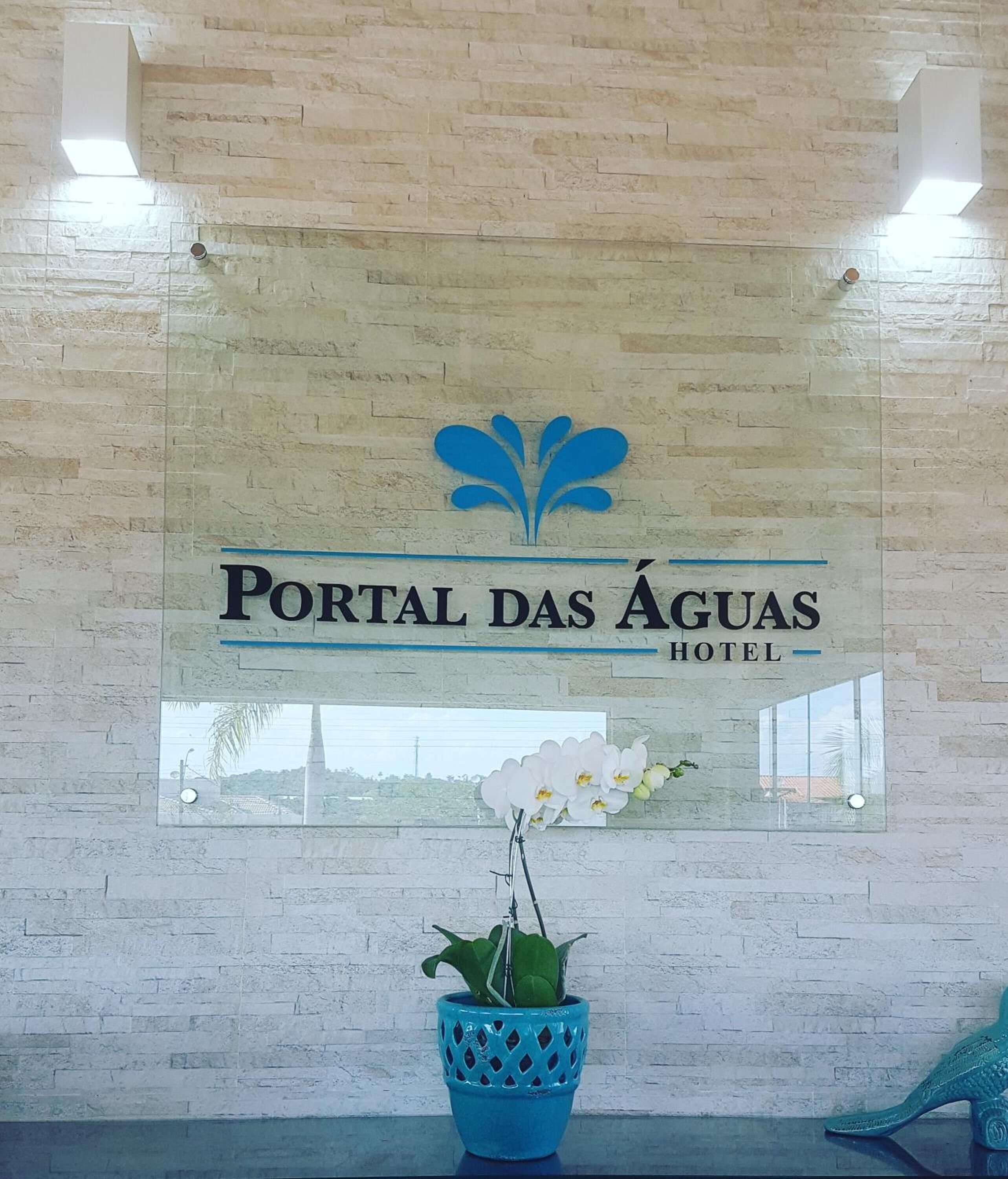Placa do Hotel Portal das Águas com logotipo, uma orquídea em vaso e uma janela refletindo uma paisagem.